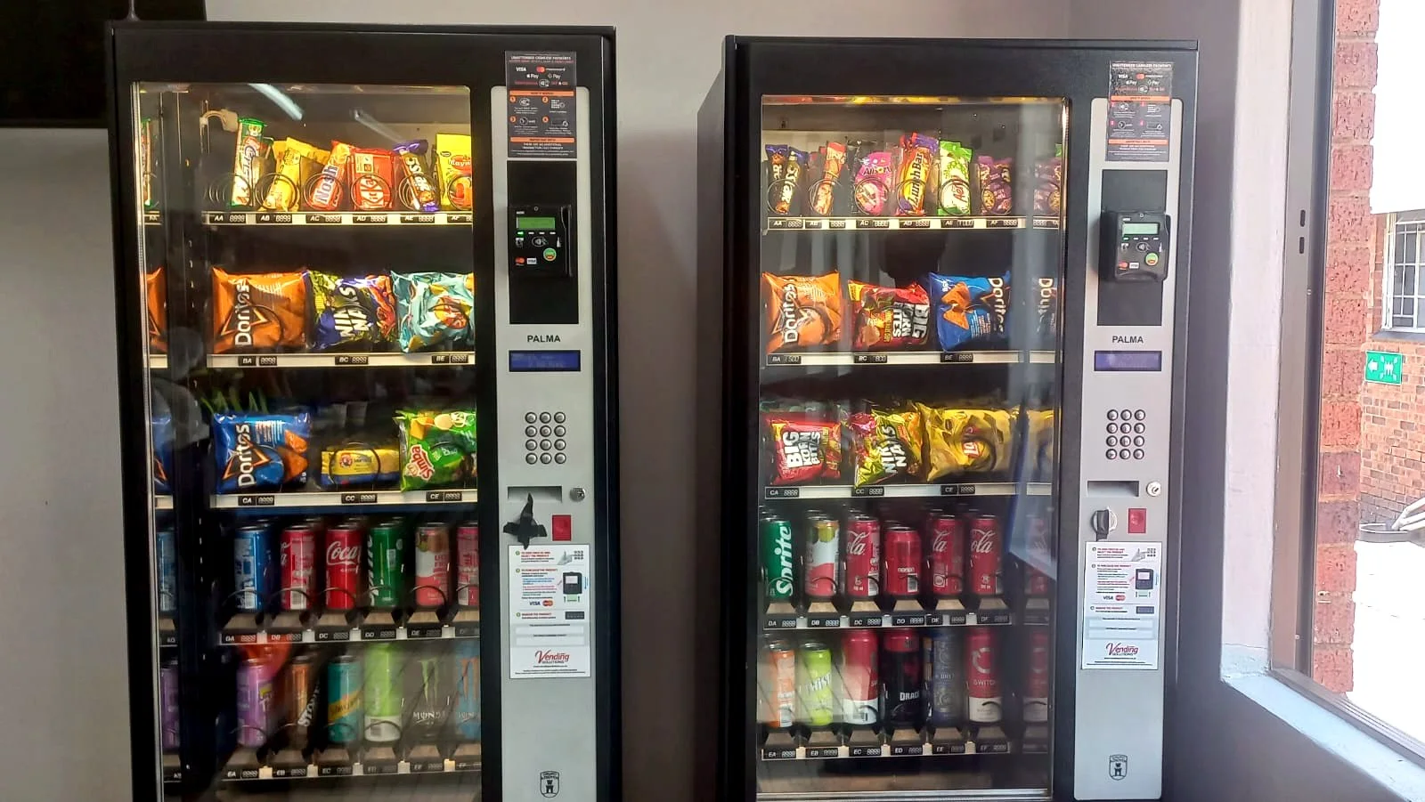 Two stocked snack and drink machines against an office window