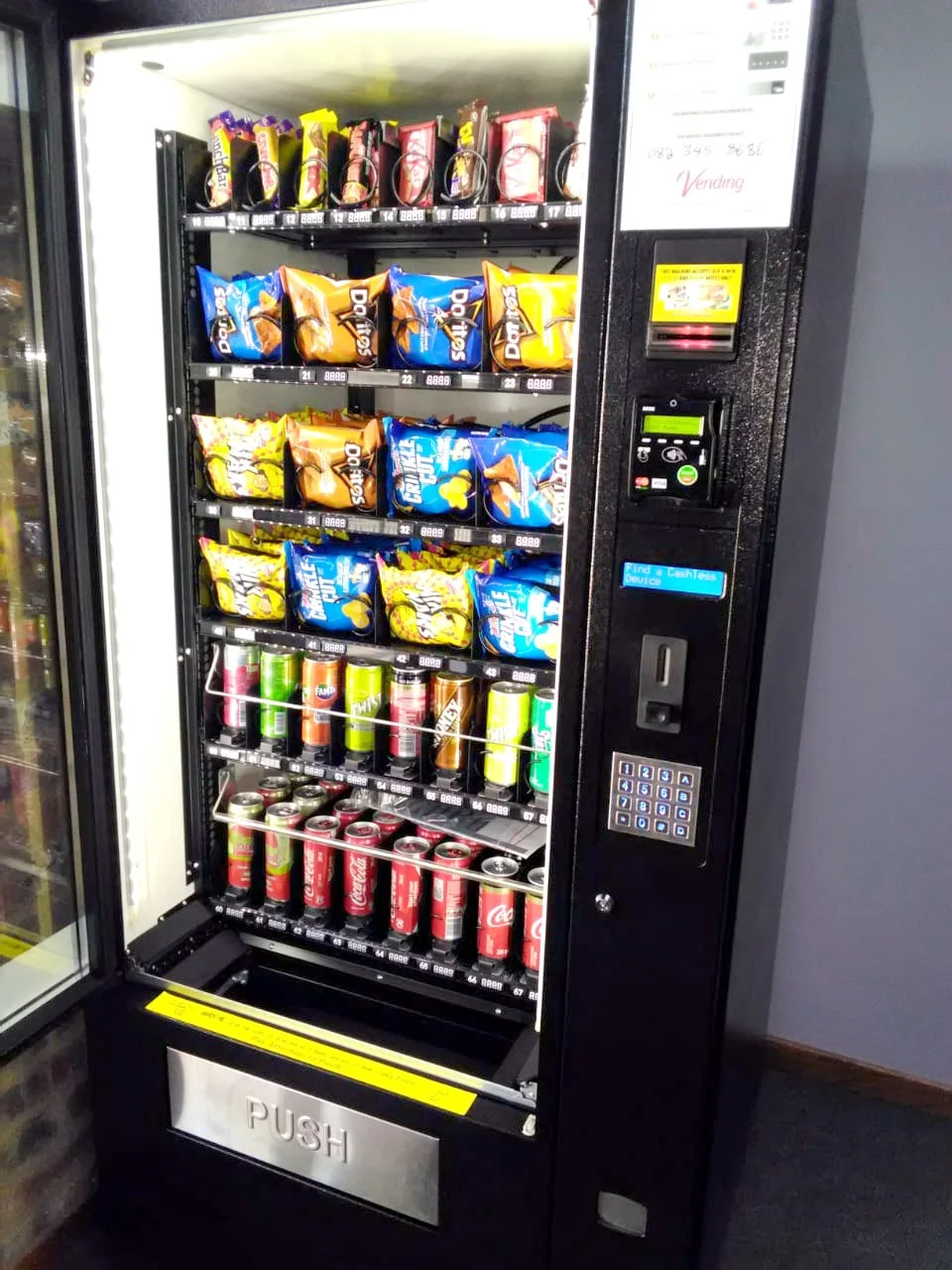 Open combination machine stocked with crisps, chocolates and cold drinks beside the keypad and card reader